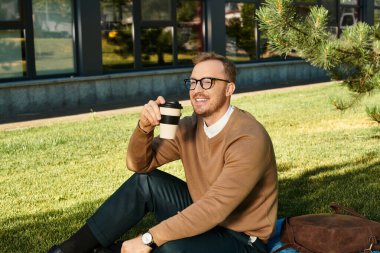 Genç bir erkek öğretmen çimenlerde otururken gülümsüyor, elinde bir kahve fincanı ve güneşin tadını çıkarıyor..