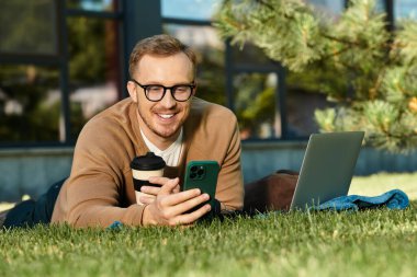 Genç adam çimlerin üzerinde dizüstü bilgisayar ve kahveyle rahatlıyor, telefonuna canlı bir şekilde odaklanıyor..