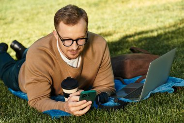 Genç bir adam yeşil bir tarlada battaniyeye uzanmış, telefonuna ve laptopuna odaklanmış, kahvesini yudumluyor..