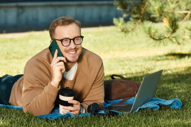 Üniversite öğretmeni çimlerin üzerinde rahatlıyor, dizüstü bilgisayar kullanıyor, telefonla konuşuyor ve kahve molasının tadını çıkarıyor..