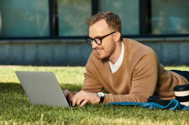 Genç bir erkek öğretmen çimlere uzanıp laptopuyla çalışıyor ve verimli bir öğleden sonranın tadını çıkarıyor..
