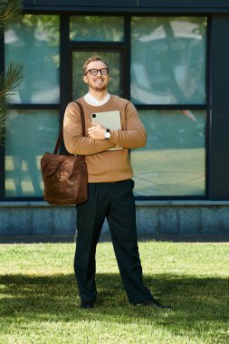 Kazaklı ve gözlüklü bir adam üniversite bahçesinde elinde bir çanta ve tabletle günün tadını çıkarıyor..