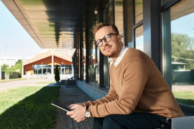 Üniversitenin dışında gözlüklü genç bir adam elinde tabletle poz veriyor ve gülümsüyor..