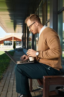 Dizüstü bilgisayarında sıradan giyinmiş genç bir adam güzel bir günde dışarıda kahve yudumluyor..