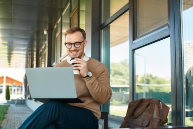 Genç bir erkek öğretmen dışarıda oturuyor, kahvesini yudumluyor ve laptopuyla meşgul oluyor..