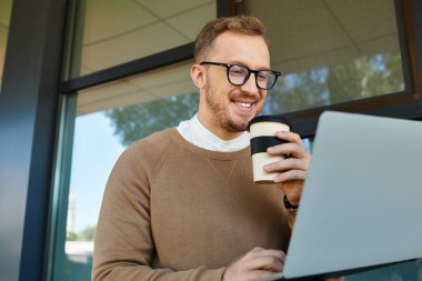 Üniversite öğretmeni dizüstü bilgisayarında çalışırken gülümsüyor, parlak bir ortamda kahvenin tadını çıkarıyor..