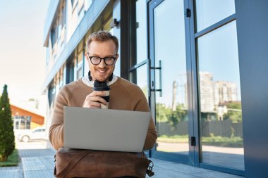 Genç adam kahveyi yudumlarken gülümser ve açık havada dizüstü bilgisayarıyla meşgul olur..