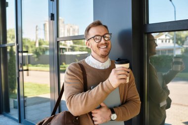 Modern bir üniversitenin önünde elinde kahve ve not defteri olan kazaklı genç bir adam duruyor..