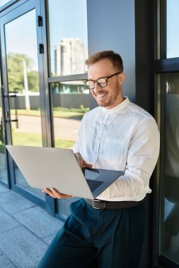 Beyaz tişörtlü ve gözlüklü genç bir adam üniversitenin önünde dizüstü bilgisayarıyla gülüyor..