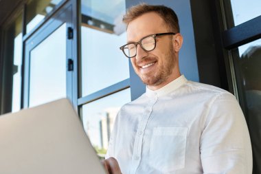 Genç bir erkek öğretmen dizüstü bilgisayarını modern bir arka plana karşı kullanırken gülümsüyor..