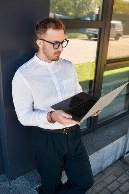 Genç bir erkek öğretmen, günün tadını çıkarırken dizüstü bilgisayarına odaklanmış bir şekilde duvara dayanıyor..