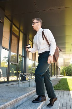 Resmi giyinmiş genç adam elinde kahve fincanı ve torbayla üniversitede merdivenlerden çıkıyor..