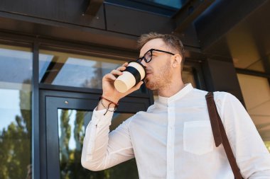Gözlüklü genç bir adam binanın dışında duruyor, kahvesini yudumluyor ve güvenle gülümsüyor..