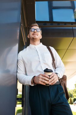 Akıllı giyinmiş genç bir adam üniversitenin önünde elinde kahve fincanıyla rahatlamış duruyor..