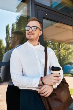 Genç bir adam cam bir girişe yaslanırken kendine güvenen bir şekilde kahve fincanıyla poz verir..