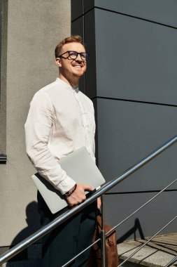 Genç adam dizüstü bilgisayarla poz veriyor, bir üniversite binasının dışındaki merdivenlerde duruyor, mutlu görünüyor..