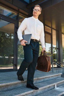 Bir üniversite öğretmeni elinde çantasıyla modern bir binadan çıkıyor, gülümsüyor..