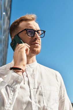 Gözlüklü genç bir adam telefonda konuşuyor, dışarıda düşünceli bir yüz ifadesi sergiliyor..