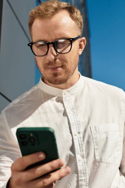 Gözlüklü genç bir adam telefonunu kontrol ederken poz veriyor..