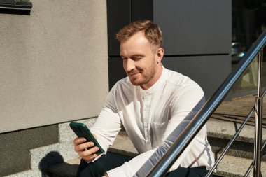 Beyaz gömlekli genç bir adam açık hava basamaklarında oturuyor, telefonuna bakarken gülümsüyor..