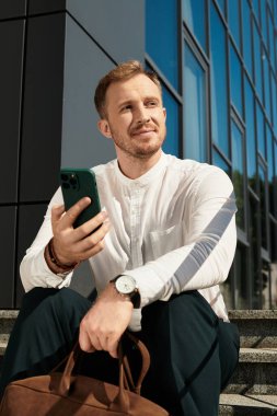 Genç bir erkek üniversite öğretmeni merdivenlerde oturuyor, gülümsüyor ve telefonunu tutuyor, güneşin tadını çıkarıyor..