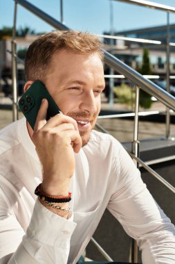 Akıllıca giyinmiş genç bir adam, üniversite ortamının dışında telefonuyla mutlu bir şekilde konuşuyor..