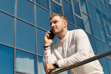 Üniversite öğretmeni olan genç bir adam, güneşli bir günde dışarıda telefonuyla konuşuyor..