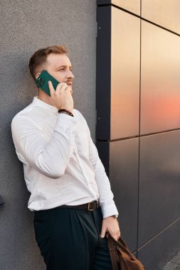 Beyaz gömlekli genç bir adam, parlak bir duvara yaslanırken telefonuyla konuşuyor..