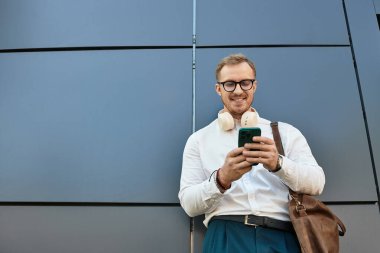 Üniversite hocası elinde çantasıyla, kulaklıklarıyla durup telefonuna bakıyor..