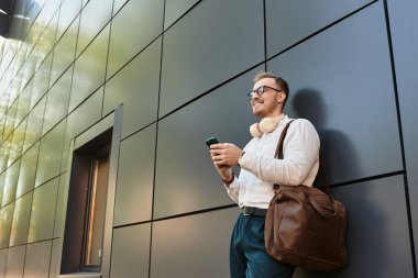 Gözlüklü genç bir adam modern bir duvara dayanıyor, gülümsüyor ve telefonunu kullanıyor..