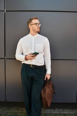 Beyaz gömlekli genç bir adam karanlık bir duvara yaslanmış, elinde telefon ve çanta, derin düşüncelere dalmış..