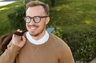 Gelişigüzel giyinen genç bir adam, arka planda yeşillikle birlikte güvenli bir şekilde dışarıda poz veriyor..