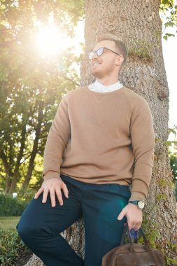 Sıradan giyinmiş genç bir adam, gün ışığında gülümserken bir ağacın yanında durur..