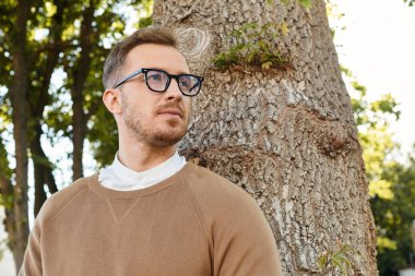 Genç bir adam, bir üniversite öğretmeni, düşüncelere dalmışken büyük bir ağacın yanında poz verir..