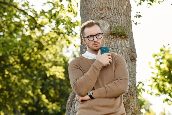 Genç bir erkek öğretmen, elinde telefonla bir ağacın yanında duruyor ve huzurlu bir ortamda dalgın dalgın bakıyor..