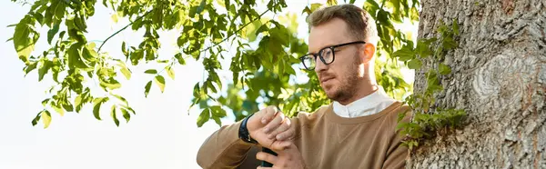 Üniversitede genç bir erkek öğretmen ağacın yanında poz veriyor, parlak güneş ışığı altında saatini kontrol ediyor..