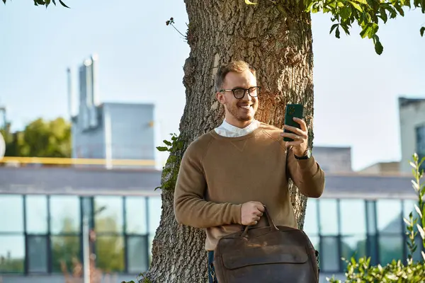 Kazaklı genç bir adam kampüsteki bir ağacın yanında telefonuna gülümsüyor..