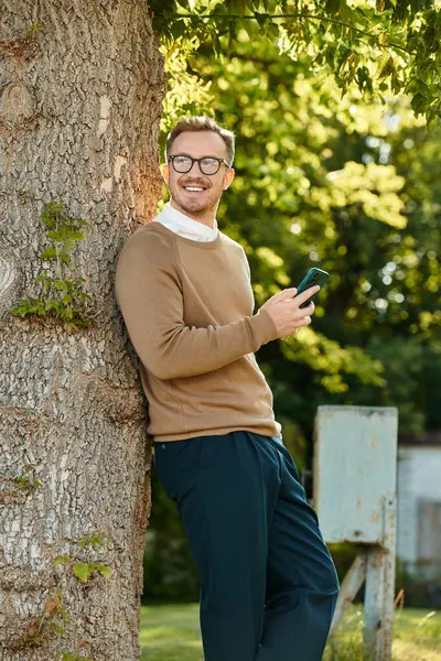 Neşeli bir üniversite öğretmeni bir ağacın yanında duruyor, telefonuyla meşgul, doğa ile çevrili..