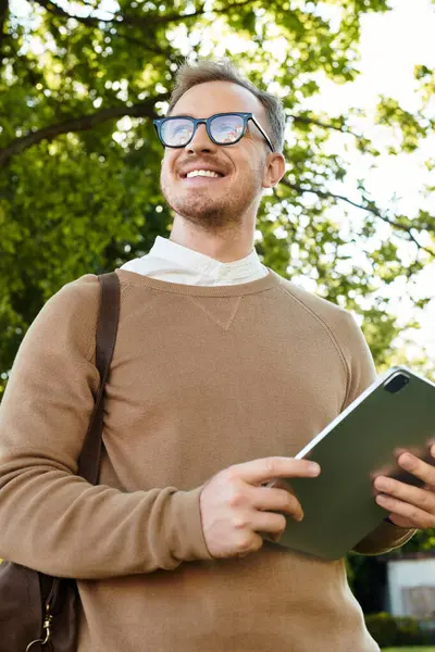 Genç adam yeşil bir açık havada elinde tabletle mutlu bir şekilde poz veriyor..
