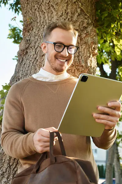 Neşeli genç bir öğretmen ağacın yanında durup gülümserken tabletle etkileşime girer..