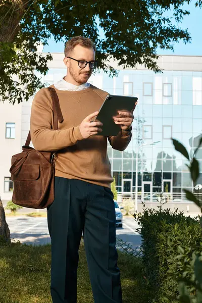 Genç bir erkek eğitmen üniversite binasının önünde poz verirken tablete bakıyor..