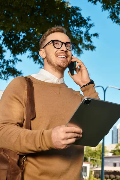 Üniversitede genç bir erkek öğretmen elinde tabletle telefonda konuşuyor..