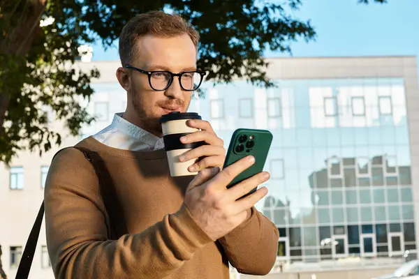 Kazak giymiş genç bir adam kahvesini yudumluyor ve kampüste telefonuna bakıyor..