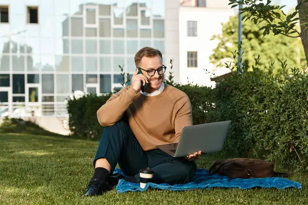 Neşeli genç bir öğretmen çim üzerinde otururken bir dizüstü bilgisayarla telefon kullanarak işe girişiyor..