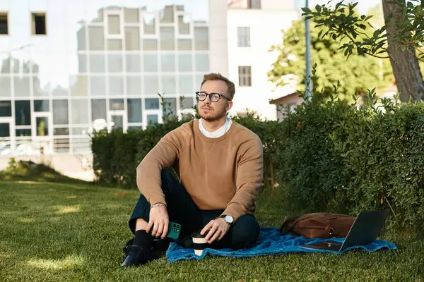 Genç bir adam yeşil bir alanda battaniyeye oturmuş kahvesini yudumluyor ve çok derin düşünüyor..