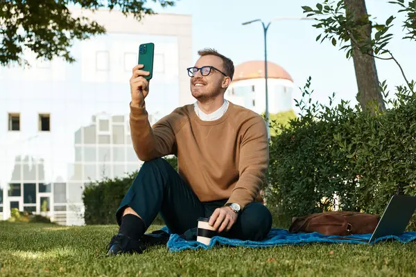 Genç bir adam çimlerin üzerinde oturur ve üniversite atmosferiyle çevrili bir şekilde selfie çekerken gülümser..