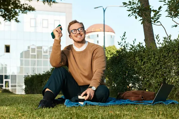 Genç bir erkek öğretmen yeşil bir açık hava ortamında rahatlarken telefon ve kahveyle gülümsüyor..
