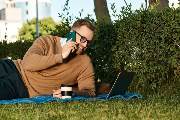 Günlük giysiler içinde genç bir adam parkta rahatlar, dizüstü bilgisayarı üzerinde çalışırken telefonla konuşur..