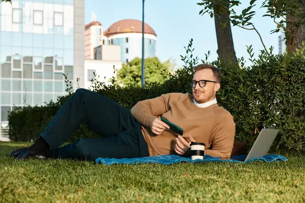 Delikanlı dışarıda çimde otururken dizüstü bilgisayar ve kahveyle rahatlamanın tadını çıkarıyor..