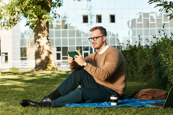 Genç bir erkek eğitimci battaniyenin üzerinde oturuyor, yeşil bir alanda akıllı telefonuyla meşgul oluyor..